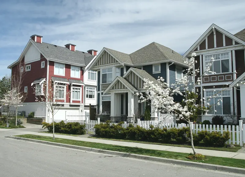 A row of modern two-story suburban townhomes in Canada featuring colorful siding in red, blue, and brown, with white fences, manicured hedges, and blossoming spring trees along a clean residential street.