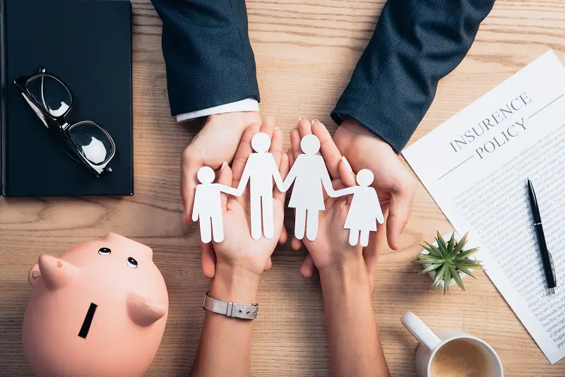 Top view of hands protecting a paper family cutout next to an insurance policy document and piggy bank, symbolizing family financial security.