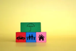 Wooden blocks featuring icons of a family, house, and car, sheltered by a top block with an umbrella icon, symbolizing comprehensive insurance protection and asset security.
